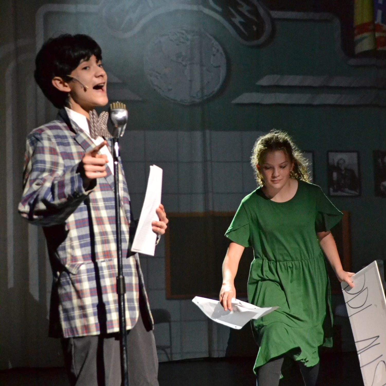 Radio boy vocalizing into a microphone while a girl is gathering papers that have been dropped
