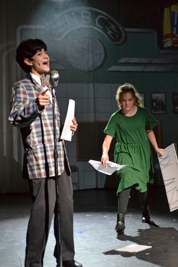 Radio boy vocalizing into a microphone while a girl is gathering papers that have been dropped