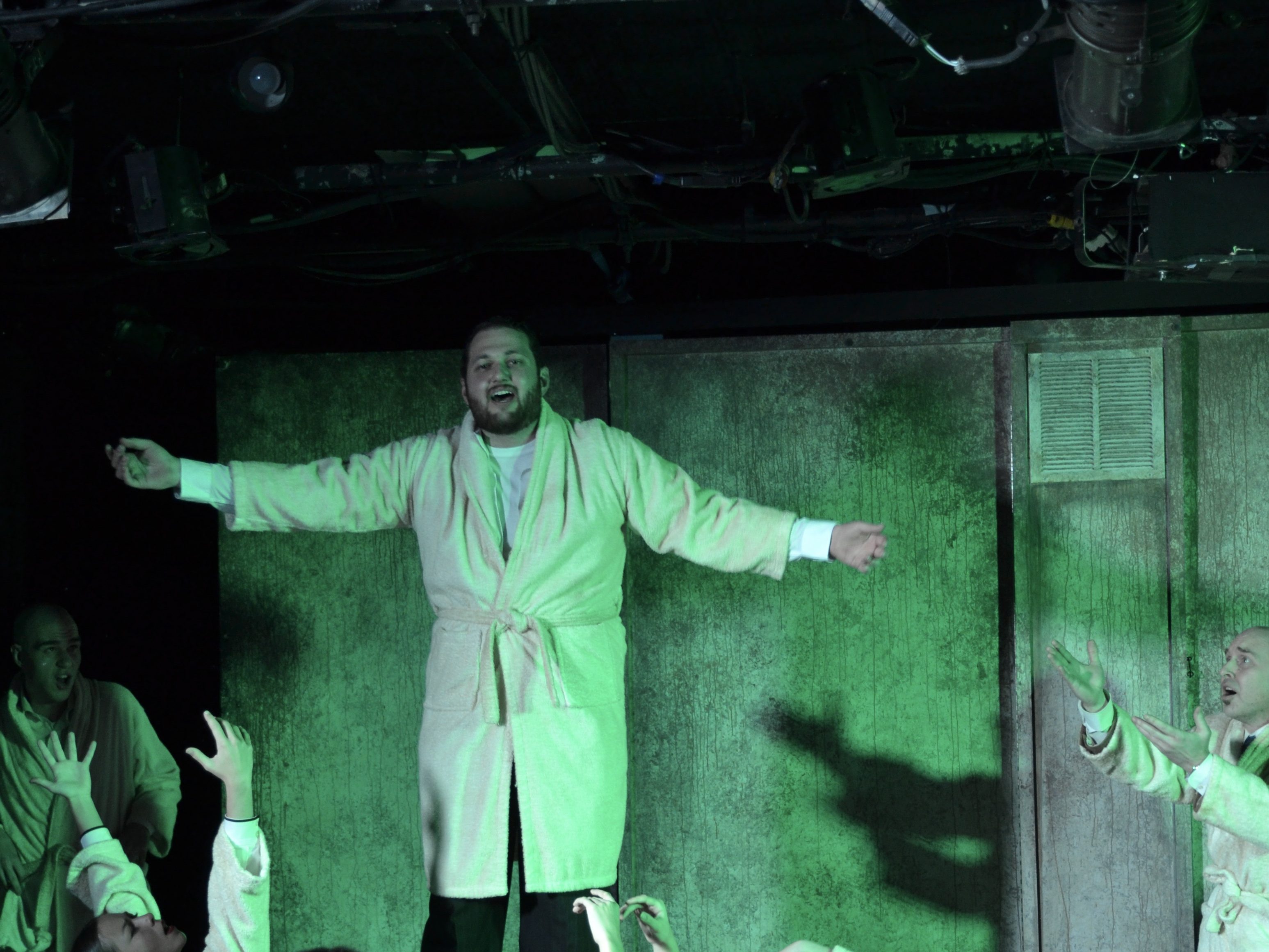 One man standing on a box, others reaching for him. All in bathrobes under spooky lighting
