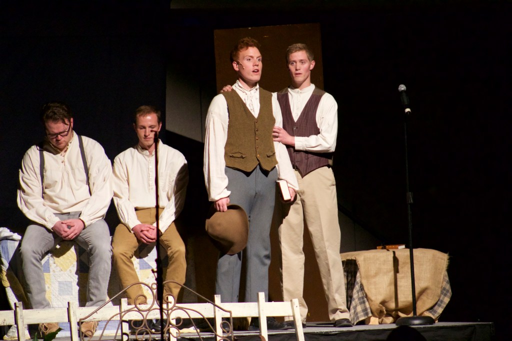 Four men, two sitting, two standing. They are dressed in Mormon pioneer garb. One of the men standing is singing.