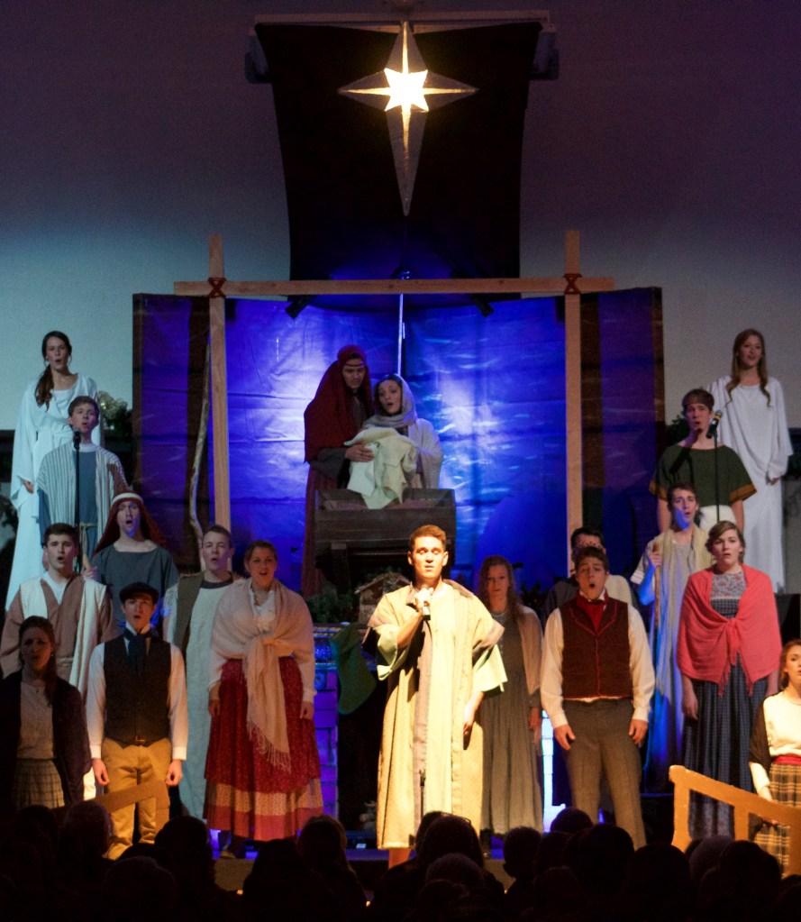 Soloist in robes holds a microphone surrounded by chorus members. Above him is a nativity scene.
